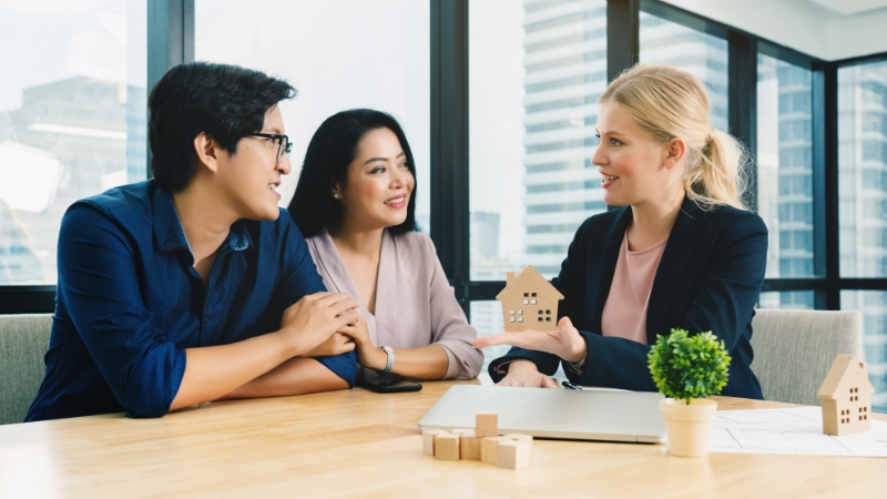 Woman explaining home equity to happy couple using wooden houses and blocks for visualization.